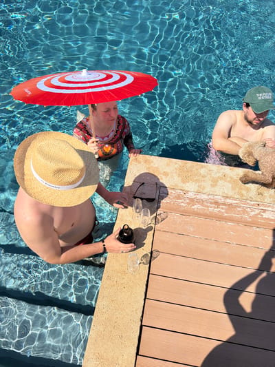 In Asciano, Italy, on a sunny day, three people are enjoying a pool. A woman is partially submerged in the water, holding a red and white striped umbrella over herself. Beside her, a man wearing a straw hat is also in the pool. A third person, a man with a green cap, is in the pool to the right, with a small dog resting on the pool edge. Champagne glasses and a bottle of alcohol are placed on the pool's edge, suggesting a celebration. The water is clear blue, with sunlight reflecting off its surface, creating ripples and shadows. The poolside is made of stone and wood, and the scene is illuminated by natural light.