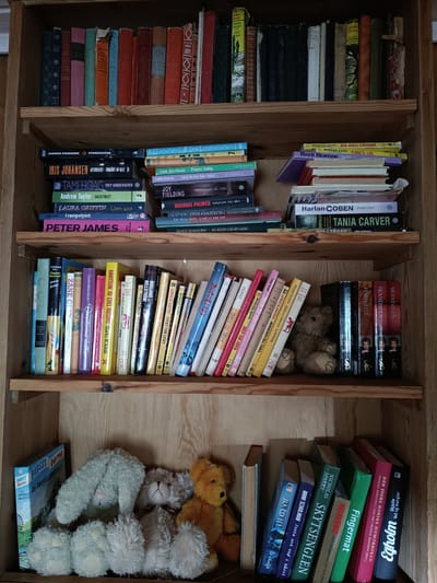 The image shows a close-up view of a wooden bookshelf filled with books and stuffed animals. The shelf is divided into three sections. The top section contains an assortment of books with colorful spines. The middle section also features a variety of books, some stacked horizontally and others vertically. A stuffed teddy bear is nestled among the books on the right side of this shelf. The bottom section of the bookshelf holds more books, a blue book with a cartoon drawing, and several stuffed animals, including a light green bunny and a gray teddy bear. The overall scene is domestic and cozy, with no clear indication of the time of day or weather.

Visible text on the book spines includes:
- "VI VOR ORD"
- "IRIS JOHANSEN"
- "TAMI HOAG"
- "ANDREW TAYLOR"
- "LAURA GRIFFIN"
- "PETER JAMES"
- "GUNNAR STAALESEN"
- "STORSESTER"
- "ATTENTATET - FORRÅDT AV ALLE"
- "DREP BUDBRINGEREEN"
- "GALGETREET"
- "UTEN SPOR"
- "IRAMPEL YSET"
- "MORGAN KANE"
- "DEN SISTE CHEYENNE"
- "KARENS LOV"
- "Berit Brænne"
- "Harlan COBEN"
- "Ringer i vann"
- "TANIA CARVER"
- "HISTORIEN OM FROKEN BERGLIOT"
- "Jorunn G. H."
- "LØRENA KICK"
- "ZANE CITY"
- "SINTA MONTEFIORE"
- "BEREKONENS DAGTA"
- "KNOTTENE AV EDEL ARGANIC"
- "CHARLES W. SCHULZ"
- "MORGAN KANE"
- "MORGAN KANE"
- "HELLSTRØM"
- "FØRLIGT"
- "SKANDALEN"
- "EGOLM"
- "Den Store Norske Humoriboken"
- "FIGERMAT"
- "SKYTSENGLEN"
- "NISSESPOOR"
- "JAG CO HU"
- "MINNA SCHULMAN"
- "Bobsey barne"
- "Og Rokkefellmysteriet"