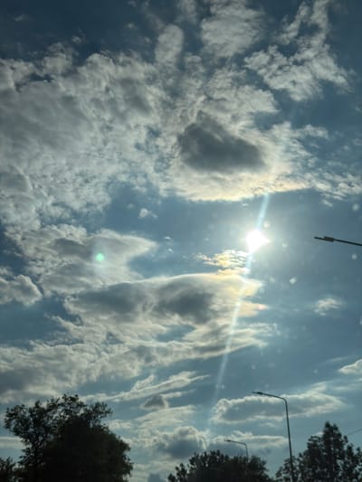 The image depicts a daytime outdoor scene dominated by a cloudy sky with the sun partially obscured. There are several streetlights visible, along with the tops of trees in the foreground. The sky is a mix of blue and grey, with the sun creating bright lens flares and a visible light beam. The overall impression is of a partially overcast but bright day. The location is identified as Teofipilka, Ukraine.