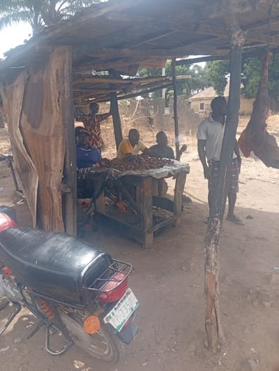 The image depicts a scene in a rural area of Assai Sabongida, Nigeria. The scene is set beneath a simple, wooden shelter, likely a makeshift stall or market stand. Four men are gathered around a table, seemingly engaged in conversation. One man is wearing a blue shirt and a red hat, another is wearing a yellow shirt, and the third wears a blue shirt. Two of the men are seated at the table, while the other two stand by the side. There is a motorcycle parked nearby, the license plate visible and reading "PP-7359," suggesting the men are waiting for a customer or are taking a break from their day. The setting is a dusty, dirt road with a small, unpaved area around the table. The scene is a typical example of rural life in Assai Sabongida, Nigeria, with a relaxed atmosphere of community and everyday interactions.