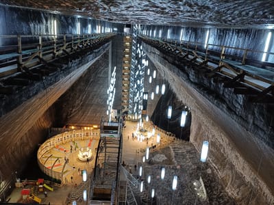 The image shows the interior of the Turda Salt Mine in Turda, Romania. It is a vast cavernous space with rough, textured walls, likely made of salt. The ceiling is also textured and appears to be illuminated by vertical, tube-shaped lights. Suspended from the ceiling are numerous hanging lights, also cylindrical and white, which illuminate the large open space.
In the center of the cavern, a multi-level metal structure, possibly an elevator or observation tower, extends upwards. Metal walkways and railings are visible along the sides of the cavern, connecting different levels and providing access.
On the lower level, a circular area is set up as an amusement attraction, resembling a mini-golf course with colorful putting greens and some playground equipment. Several people are visible in this area, including children playing on the equipment and adults observing or participating in the activities.
Further into the mine, there are more walkways, seating areas, and what appears to be a central hub with more lights and people milling about. The overall impression is of a transformed subterranean space that has been repurposed for recreational and tourist activities. The lighting creates a dramatic and otherworldly atmosphere within the salt mine.