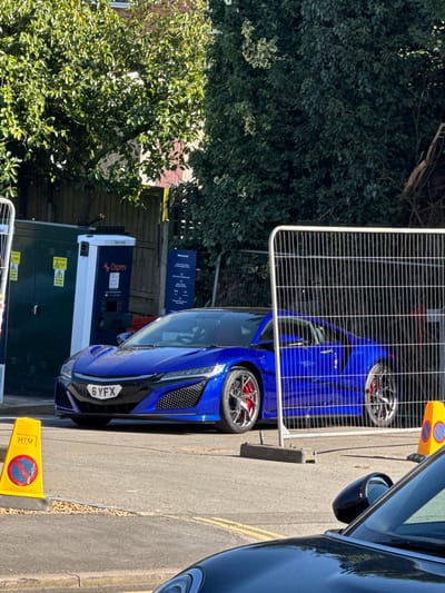 This image depicts an outdoor scene in Kenilworth, United Kingdom, on a bright, sunny day. The primary subject is a radiant blue Honda NSX (Acura NSX) sports car, bearing the UK license plate "6 YFX", parked angled slightly towards the left. The car features multi-spoke alloy wheels with prominent red brake calipers and a dark-colored roof.
To the left of the car, an electric vehicle charging station is visible. This station consists of a large, dark green electrical cabinet with yellow warning labels, and a white "Osprey" branded charging unit with "Tritium" visible on its top and a "Welcome!" sign to its right. The entire charging station area and part of the blue car are partially enclosed by temporary metal construction barriers. One barrier stands prominently in the mid-right foreground, and another is partially visible on the left.
In the lower foreground, the hood and a side mirror of a dark, likely black, vehicle are visible, suggesting the photograph was taken from within or immediately beside another car. Two yellow and orange traffic cones, marked with "HTM" and a "No Waiting" symbol, are positioned on the asphalt pavement in the lower-left and mid-right, respectively, near the barriers.
The background consists of lush green trees and bushes, with a glimpse of a reddish-brown brick building behind the foliage on the far left. The ground is an asphalt surface, and yellow double lines, characteristic of UK road markings, are faintly visible on the pavement to the right of the blue car. No people are visible in the scene, and no active charging connection is apparent on the blue car.
