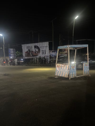 The image, taken at night in Abraka, Nigeria, showcases a street scene dominated by commercial displays. Prominently featured is a ramshackle, empty structure labeled "ABRAKA TRAFFIC STAND," its panels displaying the same text. A large banner advertising "ABRAKA ANTHEM" and promoting the release of music across all platforms hangs nearby. In the background, blurred figures and motorcycles suggest an active street life. A smaller sign mentions a "POOL PARTY" and "OFFICIAL OPENING." Streetlights illuminate the scene.