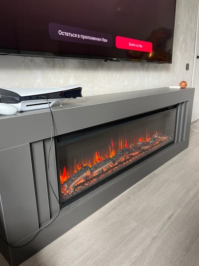 The image displays a contemporary indoor living space, featuring a large flat-screen television and a modern electric fireplace. The TV screen is active, showing a user interface with Russian text. A prominent red rectangular button reads "Выйти из Иви" (Exit from Ivi), and below it, a darker oval button says "Остаться в приложении" (Stay in the application), indicating interaction with the Ivi streaming service.

Adjacent to the television, a white PlayStation 5 console stands vertically, connected by a black cable that trails across the top of the electric fireplace. The fireplace itself is a sleek, gray unit, centrally positioned, and features an illuminated artificial flame effect with faux logs, casting warm orange and red hues.

The walls are covered with a light, textured wallpaper or paneling, and the floor is surfaced with light gray wood-look laminate planks. Above the TV, a small, spherical object resembling a baseball rests on a white wall-mounted bracket. The scene suggests a cozy, modern entertainment setup. No people are visible. The time of day is not discernible due to the indoor lighting.