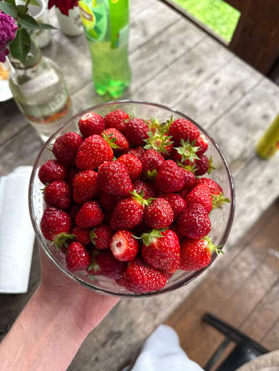 A close-up overhead shot shows a clear glass bowl filled with fresh strawberries. The bowl is held by a person's hand and arm, with visible scars on the forearm. The strawberries are plump and red, with some showing green stems and leaves. One strawberry near the bottom of the bowl has been bitten into, revealing its white interior. The bowl is resting on a weathered wooden table, with a blurred background that suggests an outdoor setting. In the background, there are partially visible items including a clear glass bottle with a label, a bright green plastic bottle, and possibly flowers. The lighting suggests it is daytime, with shadows cast on the table. The environment does not provide specific indicators of a city location.