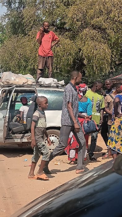 This image depicts an outdoor scene in the city of Langtang, Nigeria, on an unpaved, light-brown dirt road under daylight conditions.
In the upper-left, a bald man, approximately mid-30s to 40s, stands atop the roof of a light-colored minivan or station wagon. He is wearing a reddish-orange t-shirt with a white "QATAR" logo and dark trousers, holding a dark mobile phone to his right ear with his left hand on his hip. On the vehicle's roof, multiple irregularly shaped sacks of light-colored material, likely cargo, are secured. The vehicle's front passenger door is open, revealing a person seated inside wearing a white t-shirt and a red cap. The vehicle's rear passenger side wheel is visible.
In the foreground, a man and a young boy walk from left to right. The man, approximately mid-30s, has short dark hair and wears a patterned short-sleeve shirt (black and white design) and dark trousers with dark slip-on shoes. The boy, estimated 8-10 years old, has short dark hair and wears a striped polo shirt (brown, grey, black) and camouflage-patterned knee-length shorts with orange flip-flops.
Behind the walking individuals, a group of several people stands. One woman is visible wearing a blue patterned top, a red and white patterned wrapper, and carrying a dark blue drawstring bag. Other individuals, predominantly men, are partially obscured, including one wearing a light green t-shirt with a graphic design.
The background features dense green foliage from large trees, indicating a vegetated environment. Scattered pieces of light-colored and orange litter are visible on the ground. A dark, reflective surface, likely part of another vehicle, occupies the bottom-right corner of the frame.