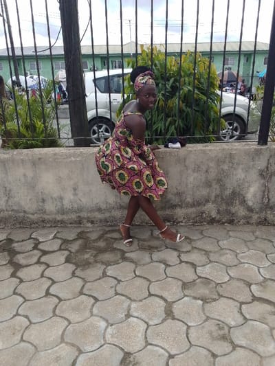 The image shows a young woman in a colorful floral dress, standing on a sidewalk in front of a concrete wall. She is wearing white high heels. The scene is in Yenagoa, Nigeria, near the coordinates (latitude: 4.931, longitude: 6.268). The background shows a car parked nearby, a building with a green wall, and lush greenery behind a metal fence. She is facing the camera but looking to her right side. The sidewalk is made of gray interlocking pavers. The weather appears to be partly cloudy. The woman's expression is relaxed and she appears to be casually posing for the photograph. She is wearing a light-colored headscarf on her head. The overall scene appears to be a typical street scene in a residential area in Yenagoa, Nigeria.