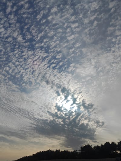 The image depicts a sky filled with patchy, cumulus clouds, with the sun partially obscured by them. The sun's rays create a bright halo effect, illuminating the surrounding clouds and casting a soft light across the scene. In the foreground, a dark silhouette of trees lines the horizon, suggesting a natural landscape. The overall lighting indicates it is likely late afternoon or early evening. There are no discernible people or specific landmarks visible that would allow for a precise location within Worthing, United Kingdom.