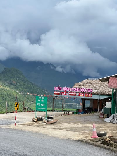 This image depicts a roadside rest stop in Lùng Tám Commune, Vietnam, under a cloudy sky. The foreground shows a paved road and a concrete area with various objects. A red and white striped pole stands next to a yellow curve warning sign. To the right, a green sign with Vietnamese text and a phone number is mounted. Further back, a thatched-roof building with a red metal roof section serves as a shop or cafe. A large pink traffic cone and a tire are visible on the concrete in the mid-ground. The background features lush green mountains, with some appearing to be partially obscured by dark clouds. The overall scene suggests a rural, mountainous location.