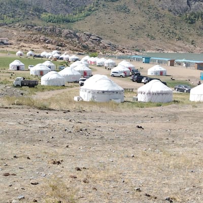The image depicts a campground with numerous white yurts situated in a dry, rocky, and grassy landscape. In the background, a hill covered with sparse vegetation and large rock formations rises. A body of water is visible further in the distance, bordered by a sandy area with several small, open-sided structures. Several vehicles, including a white truck and various cars, are parked among the yurts and on the sandy area. Some people are visible walking around the yurts and near the water. The overall scene suggests a tourist or recreational area, possibly a ger camp, in a natural, arid environment. The location in the city of Торайғыров, Kazakhstan is confirmed by the context provided.
