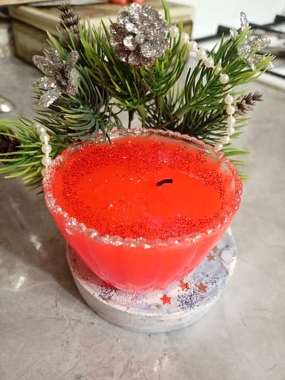 This image shows a close-up of a red, glitter-filled candle with a pine branch adorned with white beads and silver glittery ornaments. The candle is placed on a decorative base that appears to be a coaster, decorated with blue and white speckles and red stars. The background is blurred but suggests an indoor setting, possibly a kitchen counter given the visible cabinetry and stovetop. The lighting appears to be artificial, and there is no indication of the time of day or weather. The overall aesthetic suggests a festive, possibly Christmas-themed, decoration. No people are visible in the image, and there is no visible text. The image appears to be taken in Bologoe, Russia.