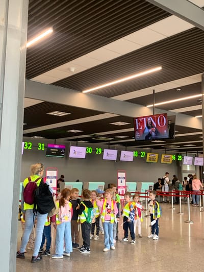 The scene is an airport terminal in Pyrzowice, Poland (latitude: 50.471, longitude: 19.072). The interior is modern with light grey walls, light beige ceiling tiles, and steel support beams. Fluorescent lights illuminate the area. A large screen displaying an advertisement for a clothing brand hangs above the check-in counters. The check-in area is lined with several counters, each with a numbered sign. There are numerous individuals standing in line at the counters. In the foreground, there is a group of children, estimated to be in kindergarten age, wearing bright colored safety vests, standing in front of the check-in counters with an adult female chaperone. The children are looking in different directions. The adults in the background are checking in for their flights. The check-in area is busy, with people moving about and bags being checked in. The atmosphere is calm and organized.