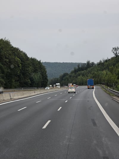 A vertical, wide-angle, eye-level shot captures a view from a moving vehicle on a multi-lane highway in Rohrbrunner Forst, Germany. The sky is overcast with a uniform light gray hue. A dense forest of dark green trees lines the left side of the highway, forming a steep embankment. On the right, the tree line is less dense and curves away into the distance. The highway itself is asphalt, marked with white lane dividers. The focus is on the road and the traffic.

Several vehicles are visible. In the foreground, on the right lane, a blue semi-truck with a white trailer is traveling away from the viewer. Ahead of it, in the left lane, a white hatchback car is also moving away. Further ahead, multiple other vehicles, including white cars and trucks, are present in various lanes. On the far left, there's a concrete barrier separating the highway from the embankment. The road surface appears wet in patches, suggesting recent rain. The overall scene depicts a typical highway drive on an overcast day.