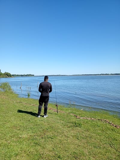A person in black clothing is standing on a grassy bank, looking out over a large body of water. The person has a fishing rod standing beside them. The sky is clear and blue. On the other side of the water is a tree-lined shore. The location is likely near the Paraná River in Corrientes, Argentina.
