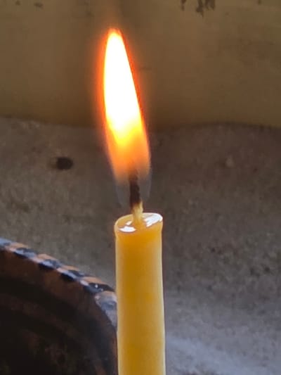 A close-up shot depicts a burning yellow candle. The flame is bright orange and yellow. The candle is placed in a dark-colored holder. The background appears to be a wall with varying textures and shades of gray and beige. The location is Balsha, Bulgaria.
