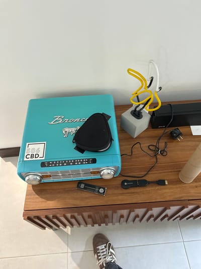 The image shows a wooden table with a teal radio on it. The radio has a sticker that says "506 CBD". The radio also has a black pouch on top of it. To the right of the radio is a black HDMI cable connected to a chromecast device. There is a gray, square, concrete lamp base with a bright yellow neon lamp in the shape of a banana on it. In the background, there is a black electronic device that is partially visible. The table is in the city of San Rafael, Costa Rica, specifically at the coordinates 9.966 latitude and -84.181 longitude. A person’s foot is visible in the bottom of the image, the person is wearing white sneakers.