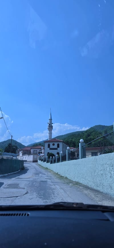The image depicts a rural village scene, identified as Melivoia, Greece, captured from inside a vehicle. The foreground shows the top edge of a dark car dashboard and a windshield wiper base. A paved road, appearing to be asphalt or concrete with visible cracks and patches, extends into the midground, sloping gently upward and curving to the left. Along the right side of the road, a light greenish-blue stucco wall runs parallel, topped by a dark metal fence with regularly spaced white spherical finials. Behind this wall is a mosque, a prominent white building with a terracotta-tiled roof and several arched windows, featuring a tall, slender minaret with a light blue conical spire. To the left of the road, a green mesh or tarp partially obscures an indistinct structure. In the background, lush, tree-covered mountains rise under a bright blue sky dotted with scattered white cumulus clouds. Faint reflections, consistent with light sources or objects inside a vehicle, are visible in the upper sky portion of the image. Two small dark specks are discernible in the sky above the minaret.