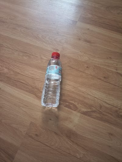 A horizontal eye-level shot shows a half-empty, transparent plastic water bottle lying on a light brown wooden floor. The bottle has a red cap and a label with blue and white text. The scene is illuminated by natural light. The location is presumed to be the city of Kubrat, Bulgaria. There are no people visible, and no specific actions or interactions are depicted in the still image.