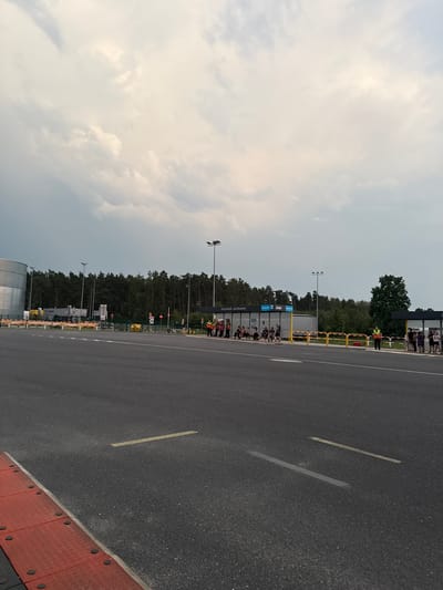 The image depicts an outdoor scene in Chociule, Poland, with a focus on a road and a bus stop area. A wide asphalt road occupies the foreground, with distinct yellow and white markings indicating lanes and the edge of the road. To the right, a red and black textured curb is visible.
In the middle ground, a bus stop structure is present, featuring a roof and clear panels. Several individuals are gathered at the bus stop, some seated on benches and others standing. A person in a bright orange vest, likely a staff member or attendant, stands near the bus stop. Further to the right, another group of people are assembled, and another individual in an orange vest is visible.
Behind the bus stop, a yellow barrier separates the waiting area from the road. Beyond this, a line of trees forms a dense forest backdrop. Several lampposts are interspersed along the bus stop area and road. In the far left of the image, a large, cylindrical metallic structure is partially visible, suggesting an industrial or storage facility. The sky above is overcast with a mix of light and dark clouds. The overall impression is that of a functional, somewhat utilitarian public transport waiting area on the outskirts of a wooded area.