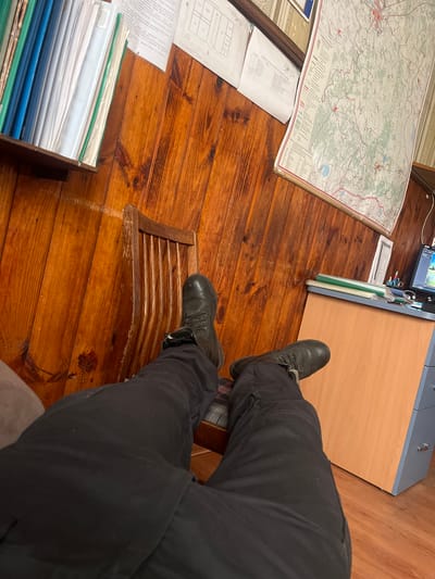 The image shows a person's legs and feet, wearing black pants and black boots. They are seated on a wooden chair with a slatted seat. The background consists of a wooden floor. On the floor, to the left, there is a large map unfurled, with text and details visible. There are also some papers and a stack of files or binders. To the right, there is a wooden desk or cabinet. The scene appears to be indoors, possibly an office or a room used for planning or research, given the presence of the map and documents. The lighting suggests it is daytime. There is text visible on the map, though it is not legible in detail. The overall impression is of someone working or studying in a room with a focus on maps and documents.