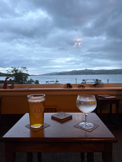 An indoor scene in Sconser, United Kingdom, captured from a slightly elevated perspective, looks out through a large window onto a body of water and distant land under an overcast sky. In the immediate foreground, a dark wooden table occupies the lower third of the frame. On the left, a pint glass containing a light amber liquid with a white foam head rests on a brown coaster, displaying a white label with "SKYE ALE." To its right, a stemmed, clear glass with a spiraled texture holds a clear beverage with ice and a lemon wedge, also resting on a brown coaster. Between the two drinks and slightly behind them, a stack of four "SKYE ALE" branded coasters is visible.

Behind the table, a wooden window sill displays various decorative items. On the left, two carved wooden duck figurines are present. On the right, a conch shell, a dark crab figurine, and a miniature crab pot are arranged. A small, dark wooden stand or table with a stack of papers or a menu is visible behind the stemmed glass.

Through the window, a wide expanse of grey water stretches towards a mountainous landmass under a heavily clouded, uniformly grey sky. A small, intense orange-yellow glow, potentially sunlight, pierces through a gap in the clouds in the upper-middle portion of the sky. Sparse foliage is visible on the immediate shoreline, and several dark structures, possibly buoys or small boat moorings, are scattered across the water. No individuals are present in the image.