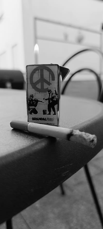 A black and white photograph shows a lit cigarette in the foreground, placed on a dark, textured surface. Behind the cigarette, a silver lighter with a flame is visible. The lighter is decorated with a stencil design of two soldiers aiming rifles towards a peace symbol, with the word "BRANDALISED" printed below. The background is blurred, suggesting an outdoor setting, possibly a table on a patio or balcony. The overall impression is one of urban imagery with a strong contrast between the symbols of conflict and peace. The location is identified as Winnenden, Germany.