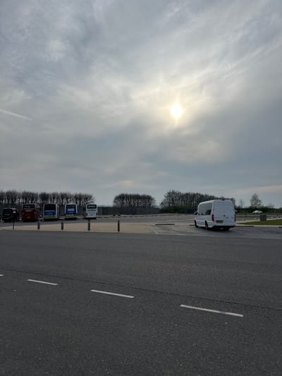 The image displays a large coach parking facility in Salisbury, United Kingdom, observed under an overcast sky with the sun appearing as a bright, circular disc in the upper right quadrant. The foreground comprises dark asphalt pavement with two parallel white dashed lines. Beyond this, a lighter-toned paved area contains numerous marked parking bays. A row of five light-colored, short bollards separates the asphalt foreground from the main parking section.
On the left side of the parking area, five large vehicles are parked sequentially. From left to right, these include a dark, partially obscured vehicle; a red coach with "COACH" visible on its rear and additional partial branding; a blue coach featuring "SALISBURY" in white text; another blue coach displaying "SWANSEA" in white text; and a white coach with "THOMAS TOURS" branding.
On the right side of the parking facility, a white Mercedes-Benz Sprinter-type van is parked in an individual bay, oriented with its rear facing the camera. Its rear doors are closed, and a UK license plate is affixed but illegible. Further in the distant background, a light-colored passenger car is faintly visible.
The horizon is delineated by a dense line of bare deciduous trees, indicative of late winter or early spring conditions. Beyond these trees, segments of open, lighter ground are discernible. No individuals are present, and no actions or interactions are depicted within the static scene.