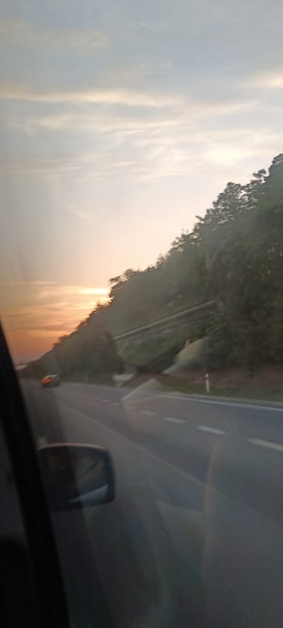 The image depicts a scenic view from a vehicle traveling on a road near Velenka, Czechia, during sunset. The road is paved and has white lane markings. To the right of the road, a steep, tree-covered hillside slopes upwards, with a guardrail visible in some areas. The sky is a pale blue with wispy clouds, tinged with orange and yellow hues from the setting sun. A single car is visible in the distance on the road, its brake lights illuminated. The foreground shows the interior of the vehicle, with a side mirror on the left and reflections on the car window. The overall impression is one of a peaceful, serene sunset drive through a natural landscape.