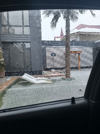 The image depicts an outdoor scene in Charvak, Uzbekistan, during ongoing heavy snowfall, captured from inside a vehicle. The foreground shows a rectangular pool or decorative pond, partially frozen or covered in granular ice and snow. A white, L-shaped floating lounge cushion, partially submerged and snow-dusted, rests within the water. To the right, a tall palm tree with a textured brown trunk and snow-laden fronds rises from a mosaic-tiled base.
In the mid-ground, a modern building with a dark grey facade and large glass windows, some displaying internal horizontal blinds, stands on the left. A grey block-tiled wall spans the background, with a grey slatted fence topping it. Against this wall, a portable basketball hoop with a blue backboard and rim is visible. A small wooden pergola structure is also integrated with the wall behind the palm tree. Further in the background, a building with a snow-covered red-tiled roof and a distinct red-pointed spire is partially visible. Snowflakes are clearly visible in the air, and all exterior surfaces show accumulation. No individuals or active interactions are present in the scene.