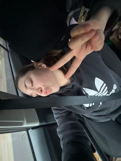 A young female subject is depicted inside a vehicle, occupying a passenger seat, secured by a dark seatbelt across her torso and lap. Her eyes are closed, and her lips are pursed in a "kiss" expression. She has light brown hair and is wearing a dark hooded sweatshirt featuring a large white Adidas trefoil logo on the front. A human hand, entering the frame from the upper right, displays a "peace" sign with the index and middle fingers extended. A gold-colored ring is visible on the ring finger of this hand. The vehicle's interior consists of dark upholstery. An indistinct, blurred exterior scene is visible through a side window on the left. No specific visual cues are present within the image to identify a precise location within Lonate Pozzolo, Italy.