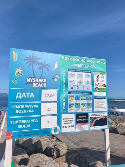 A weathered blue and white sign stands on a beach in Myskhako, Russia, against a backdrop of a bright blue sky and the sea. The sign, which appears to be a public information board for a recreational area, prominently displays "Myshako Beach" in large white letters. Sections of the sign are dedicated to providing safety information, including instructions on how to assist drowning victims, general water safety guidelines illustrated with small graphics, and a section detailing operating hours and emergency contact numbers.
Beneath the "Myshako Beach" title, temperature readings are displayed. "Temperature of air" is listed as 25 degrees, and "Temperature of water" is 21 degrees. The date "17.06" is also visible. The bottom of the sign includes legal information, mentioning "Contract for water use: OOO 'April' INN 2315180336".
To the right of the main information panel, there are additional sections with more graphics and text related to emergency procedures and evacuation plans, including a diagram of the beach area. The overall condition of the sign suggests it has been exposed to the elements for some time. The sign is mounted on a white post, which is partially obscured by large grey rocks. In the background, the sea is visible, and a few beachgoers are present near the water's edge. A white boat with "ORA" visible on its side is also in the water to the right.