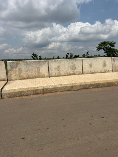 The image depicts a roadside scene, oriented vertically. On the left, a dramatic sky filled with white, puffy cumulus clouds against a pale blue backdrop dominates the view. A line of trees with dark green foliage stretches horizontally across the middle ground, creating a visual separation between the sky and the foreground.

The foreground features a concrete barrier made of rectangular blocks, weathered and discolored in places, suggesting exposure to the elements. This barrier runs parallel to a paved road on the right side of the image. The road surface is asphalt, a neutral grey-brown, with scattered dark spots that could be oil stains or debris. The overall lighting suggests it is daytime, with the sun likely casting soft light, given the even illumination of the clouds.

The scene appears to be a tranquil, everyday moment, possibly in a rural or semi-rural setting, given the presence of trees and the open sky. There are no people or vehicles visible. The concrete barrier may be part of a bridge, overpass, or retaining wall, indicating a constructed environment alongside a natural landscape. The weather seems fair but with significant cloud cover, hinting at potentially changeable conditions. The location context places this scene in Kuka, Nigeria. There is no visible text in the image.