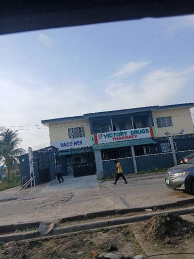 The image depicts a street scene in Ojodu, Nigeria, centered on a two-story building identified as "Victory Drugs Pharmacy" and a "Vaccines" clinic. The building features a cream-colored facade on the upper floor and a darker, possibly blue-grey, lower section with prominent signage. A metal gate with barbed wire secures the property.

Two individuals are visible on the paved area in front of the building. One person, wearing a yellow shirt and dark trousers, is walking right-to-left across the foreground. Another person, dressed in a dark shirt and trousers, is walking left-to-right near the entrance of the clinic/pharmacy.

To the right, a portion of a grey vehicle, possibly a sedan, is visible on the asphalt road. A concrete curb separates the road from a dirt shoulder where a small mound of earth is present. A palm tree is visible on the left side of the frame, behind the building's perimeter fence. The sky above is largely clear with some wispy clouds. The lighting suggests daytime conditions.
