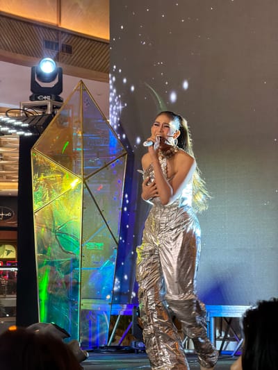 A Filipina singer, identified as Mariznette De La Cruz, is performing on stage in Cebu City, Philippines. She is wearing a silver, shimmering, one-shoulder jumpsuit and a chunky silver necklace. She is holding a microphone to her mouth and her eyes are closed in mid-performance. Behind her, a large screen displays a cosmic background with sparkling white lights. To her left, a geometric, iridescent art installation with a prism-like structure is illuminated with green and blue lights. Stage lighting is visible from the upper left of the frame. In the foreground, the backs of audience members' heads are visible.