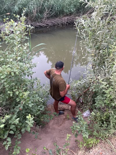 An over-the-shoulder view shows a person fishing in a narrow, murky body of water surrounded by dense vegetation. The individual, wearing a dark baseball cap, olive green t-shirt, and red shorts, stands on a dirt bank, holding a fishing rod. The fishing line extends into the water. Lush green bushes and tall reeds line both sides of the water. The water itself is calm, reflecting the muted tones of the sky and surrounding foliage. A plastic bag is visible on the ground near the person's feet. The scene appears to be a natural setting within the city of Aqsu city administration, Kazakhstan.