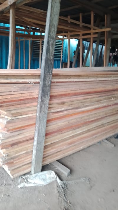 A stack of lumber rests indoors in Asubi, Nigeria. The lumber appears to be stacked on the ground with pieces of wood supporting it. A weathered, square wooden post leans against the stack. The structure around the lumber is wooden, with a corrugated metal roof. A person can be seen behind the lumber near a blue corrugated metal wall. The ground is dirt.