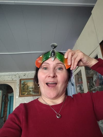 The image presents a candid, close-up portrait of a middle-aged woman captured in a selfie-style indoors, likely in her home in Сусады-Эбалак, Russia. She looks directly at the camera with a wide-eyed, open-mouthed expression of amusement, surprise, or joy. Her dark hair frames her face, and she wears a simple red long-sleeved top and a silver chain necklace with a distinctive circular pendant.
A prominent detail is the colorful, decorative object perched on her head, which she supports with her right hand. This item appears to be a bird or fish figurine, featuring translucent green and orange sections resembling wings or fins, and a grey, textured tail.
The background elements suggest a domestic setting. Above the woman, a light grey ceiling with visible panels and white strips is visible. Behind her, a wall is covered with light-colored wallpaper featuring a subtle white scroll pattern. A gold-framed landscape painting, depicting a calm scene with trees and a body of water, hangs on this wall. To her right, part of a wooden cabinet with glass doors is visible. To her left, an arched doorway or opening, adorned with blue and white striped wallpaper, can be seen. The lighting suggests it is daytime. There is no visible text in the image. The woman's playful expression is the most notable emotional cue.