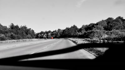 The image captures a black and white view of a curving highway in an undetermined rural area, possibly in Moldova. The road is asphalt, with white lane markings visible. On the left side of the road, a densely wooded area with trees forms a natural barrier. On the right side, a similar treeline is present, with some dry grass and a low barrier structure. In the distance, a red vehicle is traveling on the highway, and a black vehicle is also visible on the road ahead. The foreground of the image is dominated by a large, dark shadow, likely cast by a vehicle or structure from which the photograph was taken. The sky above is clear and bright. There are no people clearly visible in the image.