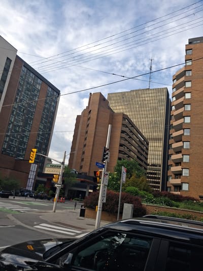 This is a street-level photograph taken in Ottawa, Canada, focusing on an urban intersection with multiple buildings and street infrastructure under a cloudy sky. The foreground is dominated by the hood and windshield of a black vehicle, obscuring the immediate street surface.
To the left, a multi-story building with a brick and glass facade is angled into the frame. It features a prominent yellow traffic light on a pole. Adjacent to this building, a pedestrian crossing with white stripes is visible on the asphalt road, with green painted lanes indicating a bike path.
In the midground, several more buildings rise up. A tall, thin, brick building with recessed balconies and numerous windows is on the right. To its left, a wider, darker brick building with a stepped facade is prominent. Further back and slightly to the left is a very tall building with a golden-tinted glass facade, topped with a radio antenna.
Street signs are visible, including a blue street sign reading "rue Queen St" with associated numbers and a red traffic signal. Several traffic cones are scattered near the curb in front of an entrance to a building. Greenery, including trees and bushes, is planted along the side of some buildings, creating a contrast with the urban architecture. The sky is partly cloudy, with white and grey clouds against a pale blue backdrop. Power lines are strung across the upper portion of the image.
