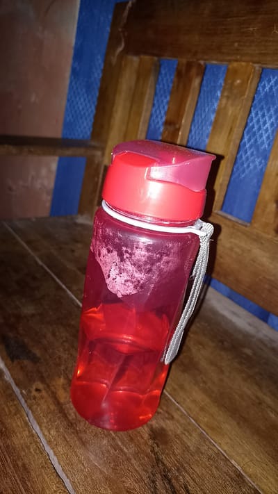 A red plastic water bottle with a red cap is placed on a wooden surface. The bottle is partially filled with a red liquid and has a white strap attached to its neck. The background consists of a wooden chair with a blue fabric covering. The image appears to have been taken indoors, likely in a residential setting in Sedayu, Indonesia, given the rustic wooden furniture and simple background.