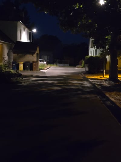 A nighttime view of a street in Kostelec na Hané, Czechia, illuminated by a streetlamp and lights from buildings. On the left, a white building with a single window is visible, adjacent to a brick structure with a slanted roof. In front of the brick structure are four trash bins: one blue, one yellow, one black, and one brown. A white car is parked further down the street on the right side. Trees with dark foliage frame the scene, casting shadows on the asphalt road. The overall impression is of a quiet, dimly lit residential or commercial street after dark.