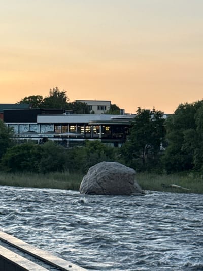 The image depicts a body of water with choppy waves, likely a river or large lake, in the foreground. On the far bank, there is lush green vegetation and grass. Behind the vegetation, a large, grey boulder is visible. Further inland, a modern building complex with a dark roof and white facade is situated among trees. Several other buildings, including a taller structure with visible windows, are scattered in the background. The sky above is a soft gradient of orange and yellow, suggesting either sunrise or sunset. A wooden walkway or platform is partially visible in the lower left corner of the image. There are no people or animals visible in the scene. The location is identified as Askhazan, Russia, implying an urban or semi-urban setting.