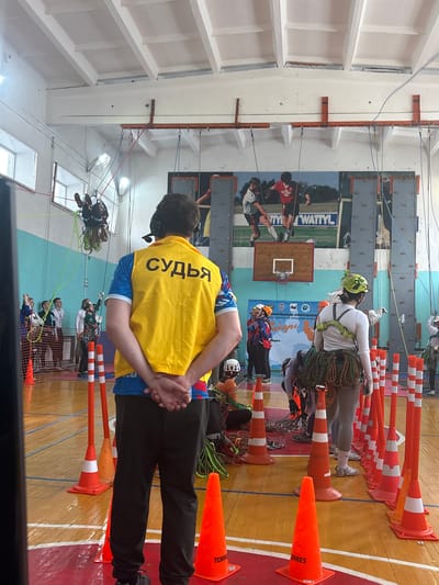 This image captures an active indoor rope competition or training event within a sports hall in Tayshet, Russia.
In the foreground, a male individual stands with his back to the camera, wearing a blue and white patterned shirt, black pants, black shoes, and a yellow vest with "СУДЬЯ" (JUDGE) printed in black Cyrillic letters. He wears an over-ear headset with a microphone and has his hands clasped behind his back. Around him and extending into the midground, numerous orange traffic cones, some with white reflective bands and "TORRES" branding, are arranged on the polished wooden gymnasium floor, which features orange and blue painted lines.
In the midground, to the right, a female participant stands facing away from the camera, wearing a white long-sleeved shirt, grey leggings, a green climbing harness, and a green helmet with decorative elements. Multiple coiled ropes of varying colors are attached to her harness, draped around her lower back. To the left midground, an individual is suspended upside down from multiple ropes near the ceiling, clad in dark clothing and a helmet. Below this suspended figure, other individuals, some wearing helmets, stand on the floor near a light blue and white wall, observing or waiting.
The background wall features a large mural depicting two male soccer players, with the text "WATTYL" visible. Below this mural, a wooden basketball backboard and hoop are mounted. To the left of the basketball hoop, an event banner with logos and Cyrillic text, including "ТУРИЗМ" (Tourism) and "ТАЙШЕТ" (Tayshet), is displayed. To the far right, sections of a vertical climbing wall with various handholds are visible. An extensive network of colored ropes (green, blue, red) descends from orange metal beams and anchor points affixed to the upper walls and ceiling structure. The ceiling is white with exposed support beams and fluorescent lighting. Large windows are visible on the upper left wall. The scene is brightly lit, indicating a well-equipped indoor facility for technical rope activities.