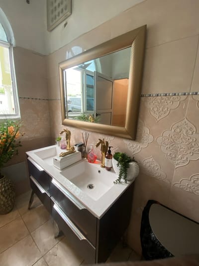 The image shows a bathroom vanity in a home located in Buea, Cameroon. It is a modern bathroom with a double sink, a mirror with a gold frame, and several decorative items, including a white vase with green plants, a gold soap dispenser, and a gold faucet. The vanity has a black wooden cabinet with three drawers. The walls of the bathroom are tiled in a light beige color with decorative white floral patterns. The floor is also tiled in a similar shade of beige. There is a small, black object on the floor in the lower right corner of the image. The location of the bathroom is based on the coordinates provided. A window with blinds can be seen in the background, but the rest of the room is not visible.