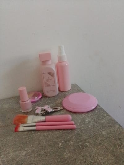 A flat lay image shows a collection of pink cosmetic items arranged on a textured, grey surface, possibly a countertop or shelf. The items include two pink spray bottles, one taller than the other, a pink bottle with a gold label and stopper, a pink nail polish bottle, a pink compact, a pink hair clip with a bow, and three makeup brushes with pink handles and colored bristles. The background is a plain, light-colored wall. The scene is indoors. There are no people present. The location is identified as Nor Geghi, Armenia.