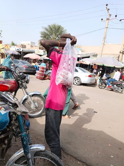 A man in a pink shirt and knit cap stands in a street market, carrying a plastic bag and containers of what appear to be strawberries. He supports the bag on his head with his hands. Several motorcycles are parked nearby, and cars and pedestrians fill the background. The market is shaded by umbrellas and includes various stalls and buildings. Overhead power lines extend from a utility pole. The street is paved and shows signs of wear.