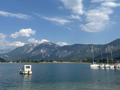 The image depicts a serene lakeside scene in Calceranica al Lago, Italy. In the foreground, the calm, blue water of the lake stretches towards a distant shoreline lined with trees and buildings. A small white boat with a canvas cover is moored in the middle of the lake, with a yellow buoy attached. Further in the distance, several sailboats are docked, their masts reaching towards the sky. The backdrop is dominated by a majestic mountain range, its rugged peaks partially obscured by scattered white clouds against a clear blue sky. The overall atmosphere is peaceful and picturesque, suggesting a pleasant day in a scenic natural setting.