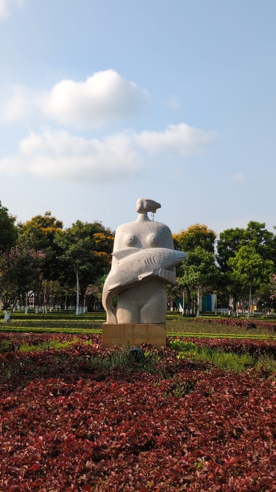 A statue of a nude woman embracing a fish stands in a park in Phan Rang – Tháp Chàm, Vietnam. The statue is made of light-colored stone and is positioned in a bed of red plants. Behind the statue, there are trees, a green strip of lawn, and metal barriers. The sky is visible in the background, with a few clouds.