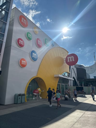 Here is a description of the image.
The image shows a building with an M&M's logo in Lake Buena Vista, Florida, United States. The building has a white facade with several large, colorful M&M's logos on it. The building is shaped like an M&M's candy and has a large yellow section with a red M&M's logo on top. There are people walking on the sidewalk in front of the building. The sky is blue with some clouds, and the sun is shining brightly. There is a sign that says "WELCOME TO M&M'S." Another sign says "WORKS SPRINGS." The image is rotated slightly to the left. There are trash cans visible.