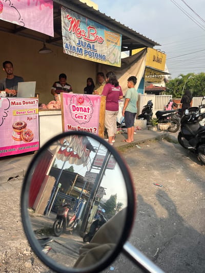 The image depicts an outdoor market stall in Sugih Waras, Indonesia, selling "Mas Bro Jual Ayam Potong" (Mas Bro Sells Cut Chicken) and "Donat Gen Z" (Gen Z Donuts). A man in a dark t-shirt stands behind a counter, appearing to be a vendor. To his right, another man in a striped shirt is preparing meat, with raw chicken visible. Further to the right, a woman wearing a dark hijab stands next to a man in a light-colored shirt. In front of the stall, a young boy in a green t-shirt and shorts stands with a young boy in a purple t-shirt and beige pants. The stall features pink banners advertising donuts, with images of donuts and text in Indonesian. One banner reads "Mau Donat?" (Want Donuts?). Another banner on the right reads "Donat Gen Z Aja...!" (Just Gen Z Donuts!). The background shows a yellow shipping container with "JA. BAGS" written on it and some residential buildings and trees. Several motorcycles are parked nearby. The foreground of the image is dominated by the reflection of the scene in a motorcycle's rearview mirror, showing a red scooter, the street, and the shadows of people. The overall scene suggests a bustling local market environment.